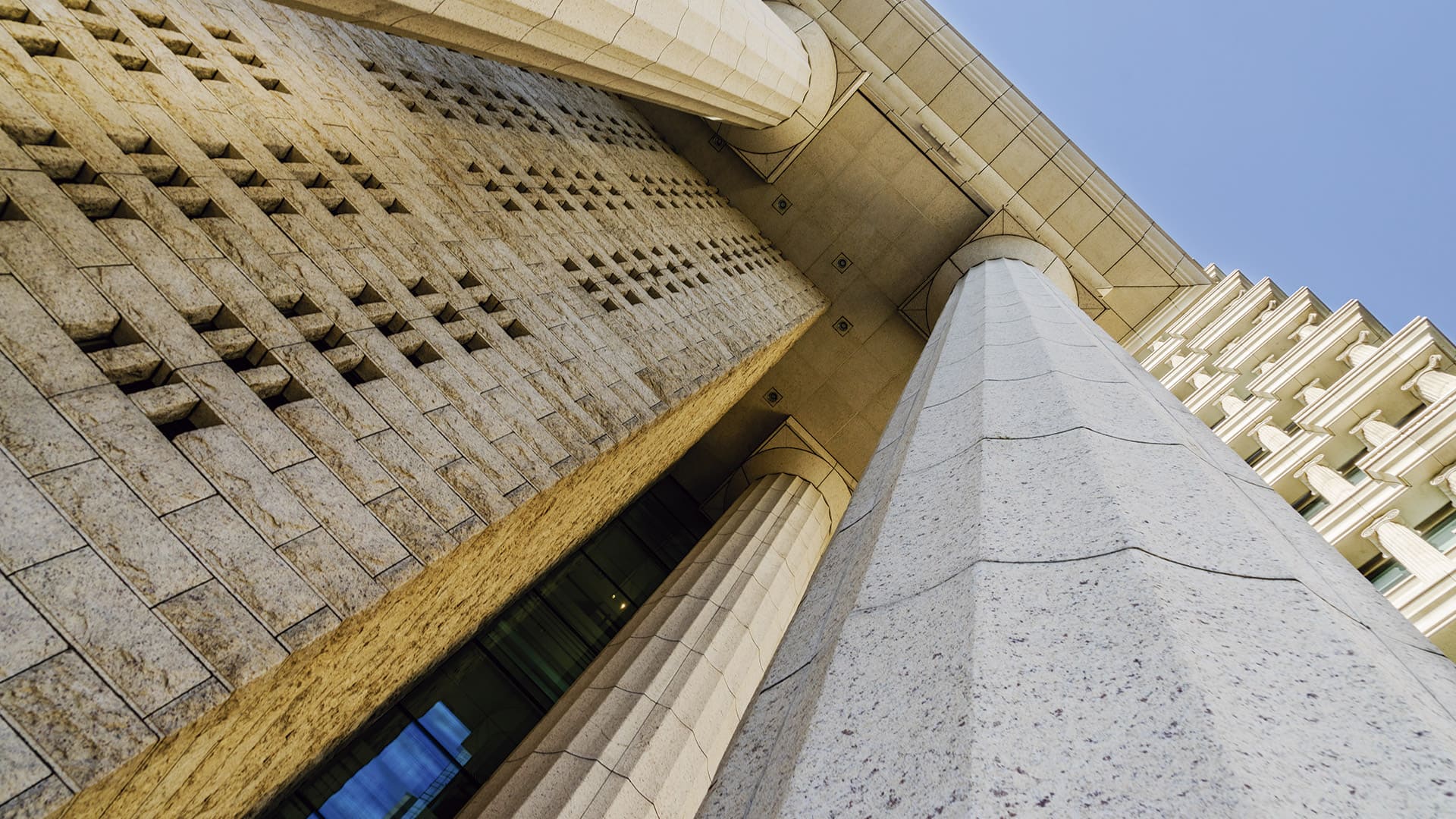 Imagem de uma imponente fachada de um edifício com colunas clássicas e detalhes arquitetônicos em pedra. A perspectiva da foto, tirada de baixo para cima, destaca a grandiosidade das colunas e a textura das paredes, com o céu claro ao fundo.